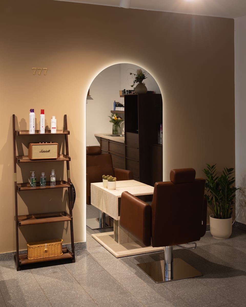 Ein Friseurstuhl vor dem beleuchteten Bogen, daneben ein Holzregal mit Pflegeprodukten